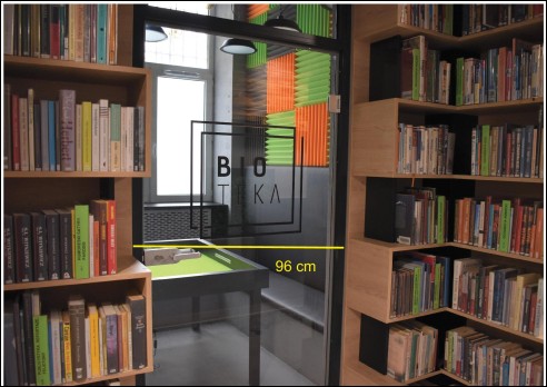 Po lewej i prawej stronie regały z książkami, na środku przeszklone drzwi z napisem BIOTEKA.