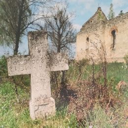 Stary kamienny krzyż wśród trawy w tle ruina kaplicy